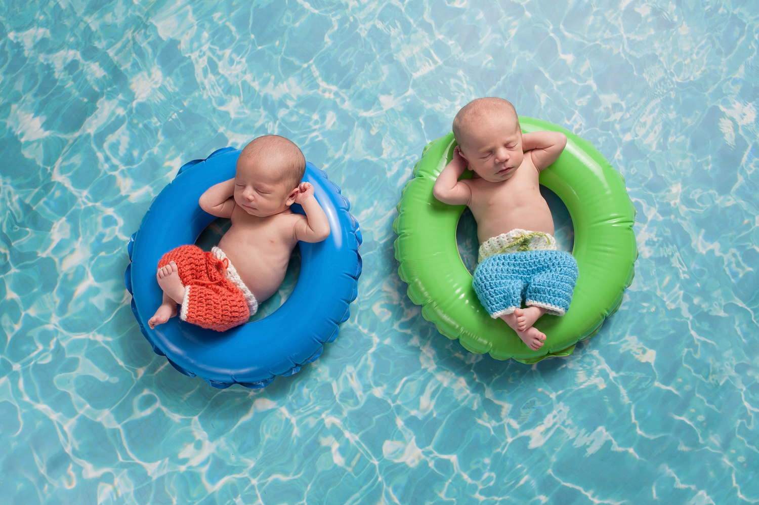Four week old twin, newborn, baby boys wearing crocheted board shorts and sleeping on a tiny inflatable swim rings. Shot in the studio on a background to look like swimming pool water.